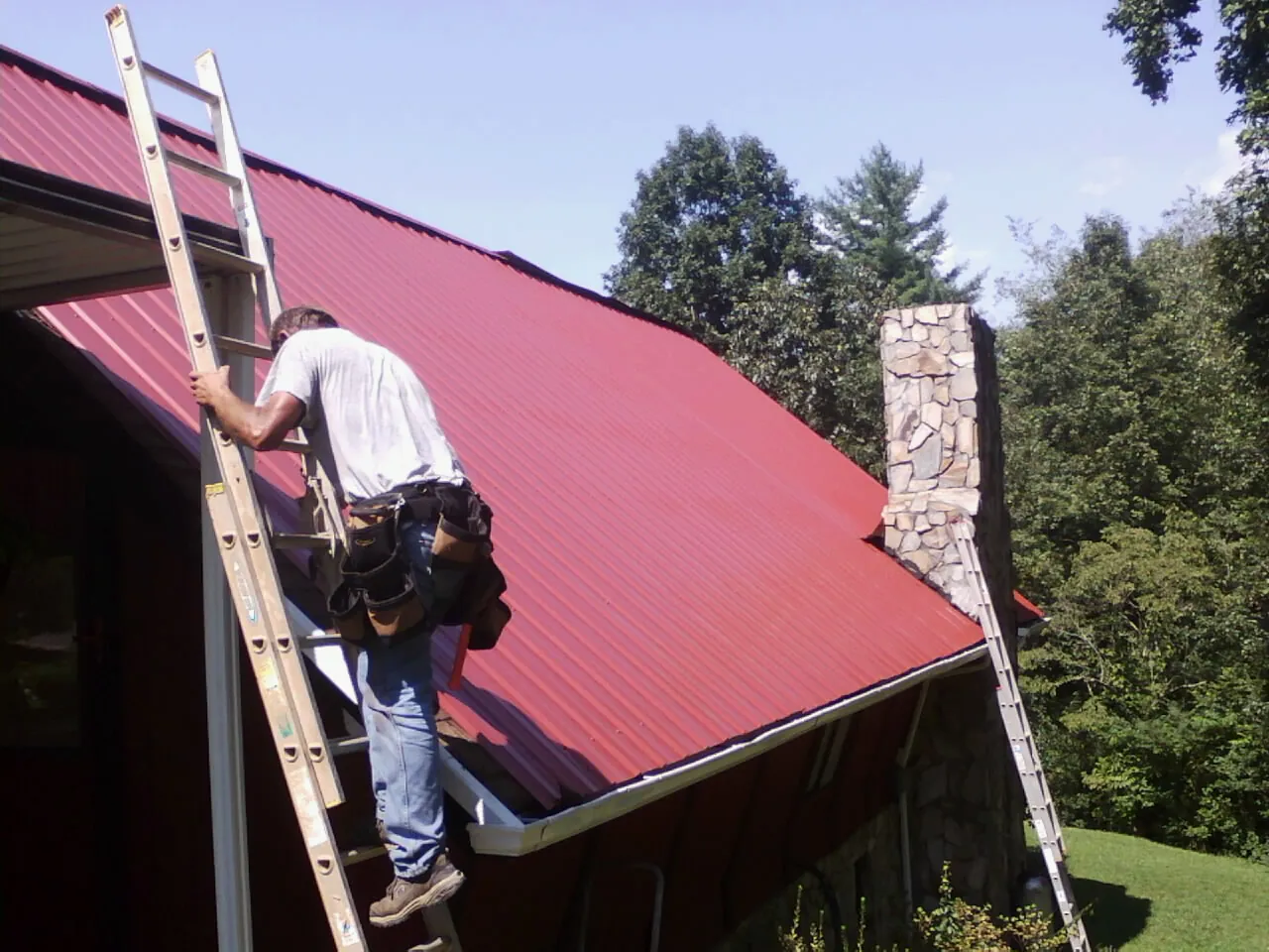 Roofer on a ladder beside a red metal roof and stone chimney — Soffit & Fascia Repair by Roofing Brothers in Guymon